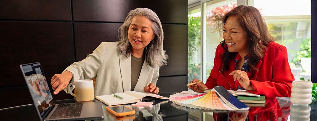 Two women laughing together over a laptop, a notebook and a fan of colour swatches while planning a project